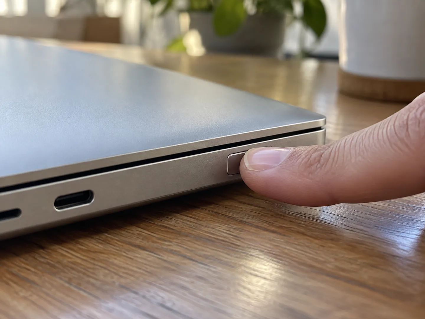 A fingertip resting on the fingerprint sensor of a closed laptop on a wooden desk